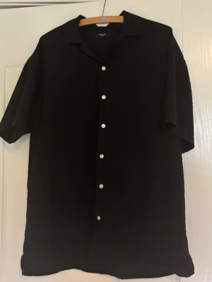 Svart kortärmad skjorta Jack & Jones - Stilren svart skjorta från Jack & Jones Premium med korta ärmar och klassisk krage. Skjortan har knappar framtill och en avslappnad passform, perfekt för en clean look. Materialet känns mjukt och svalt, riktigt nice till sommaren.