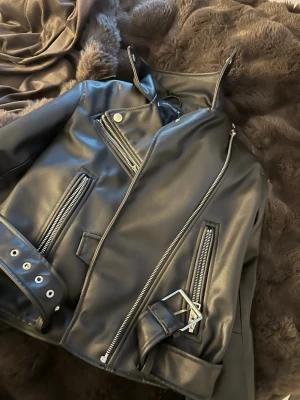 Svart biker skinnjacka med bälte - Snygg svart skinnjacka med silvriga dragkedjor, bälte i midjan. Jackan har flera fickor med dragkedja och coola detaljer som ger en edgy look. Använd 1 gång!