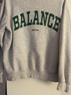 Grå sweatshirt BALANCE tryck - Snygg grå sweatshirt med mörkgrönt BALANCE-tryck på bröstet och texten WITHIN under. Materialet känns mjukt och bekvämt, troligen bomullsblandning.