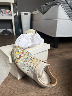 Maison Margiela sneakers med färgstänk - Unika sneakers från Maison Margiela i vitt läder med färgstänk i gult, rött, blått och grönt. Klassisk låg modell med beige sula och vita skosnören. Insidan är fodrad i ljusbrunt läder för extra komfort. Perfekta för dig som vill sticka ut. Köpta helt nya från merchsweden, digitala kvitton finns så skriv om du vill ha bevis på att de är köpta där ifrån, bra skick bara smutsiga vilket går att tvätta bort.