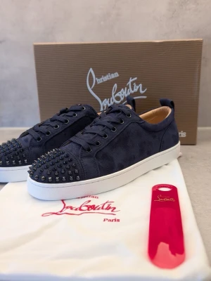 Mörkblå sneakers från Louboutin - Snygga mörkblå skor i mocka från Christian Louboutin med svarta nitar på tån och vit sula. Skorna har klassisk låg profil, svarta skosnören och exklusiv känsla. Perfekta för dig som vill sticka ut med en edgy detalj.