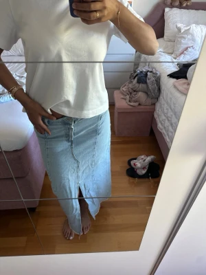 Ljusblå lång jeanskjol med slits - Snygg ljusblå lång jeanskjol i klassisk denim med slits framtill och rå kant nertill. Kjolen har en bakficka och enkel design som passar till många olika stilar. Färgen är som bilderna nerjaga bär plagget! Etikett av men har för mig från H&M