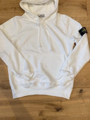 Vit hoodie från Stone Island - Vit hoodie från Stone Island med klassisk magficka och justerbar huva med dragsko. Ikonisk svart logotyp-patch på vänster ärm. Tillverkad i mjuk bomull för en skön och avslappnad stil.
