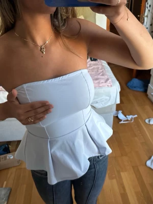 Ljusblå axelbandslös peplum tubtopp - Snygg ljusblå  axelbandslös tubtopp med peplumvolang nertill. Toppen har en figurnära passform upptill och markerad midja som ger en feminin siluett. Perfekt att styla med jeans eller kjol för en trendig look. Den ser väldigt vit ut i den två första bilderna men lite schystare syns den på sängen
