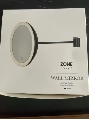 Svart sminkspegel från Zone Denmark - Rund väggmonterad sminkspegel i svart från Zone Denmark med 5x förstoring och tre olika ljusinställningar. Spegeln har en stilren design med LED-belysning runt kanten och en justerbar arm för flexibel placering. Perfekt för detaljerad makeup och hudvård.