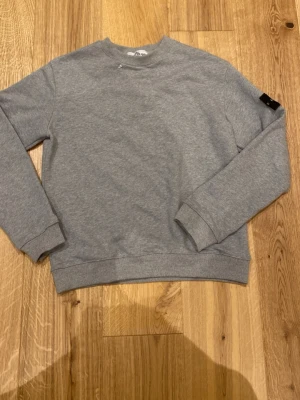 Grå sweatshirt från Stone Island - Snygg grå sweatshirt från Stone Island med klassisk rund halsringning och ribbade muddar. Tröjan har en enkel och stilren design med en liten svart logotyp på ärmen. Perfekt för en avslappnad och trendig look.