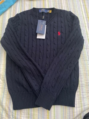 Mörkblå kabelstickad tröja Polo Ralph Lauren - Mörkblå kabelstickad tröja från Polo Ralph Lauren med rund halsringning och röd broderad logga på bröstet. Tröjan har ribbade muddar vid ärmslut och nederkant. Perfekt för lager-på-lager och klassisk stil.