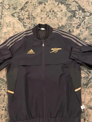 Svart Adidas Arsenal vindjacka - Snygg svart vindjacka från Adidas x Arsenal med guldiga detaljer och loggor. Jackan har dragkedja framtill, ribbade muddar och grå ränder på axlarna. Perfekt för dig som vill ha sportig stil med klubbkänsla.