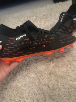 Puma Future svarta och orange fotbollsskor - Snygga fotbollsskor från Puma Future-serien i svart med orange detaljer och orange sula med fasta dobbar. Skorna har snörning och stickad ovandel för bra passform och kontroll på planen. Perfekta för dig som vill sticka ut på fotbollsplanen.