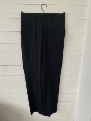 Svarta kostymbyxor från Hejco - Snygga svarta kostymbyxor från Hejco med klassisk rak passform och pressveck. Byxorna har hällor för skärp och stängs med knapp och dragkedja. Perfekta för en stilren look och passar till många olika outfits.
