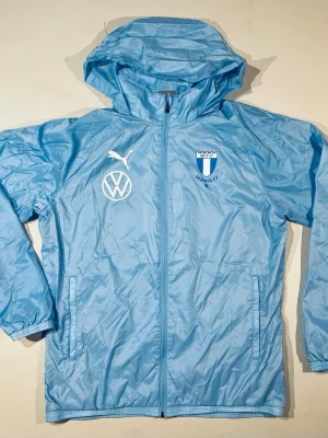Spelar jacka MFF - ANVÄND AV SPELARE Ljusblå vindjacka från Puma / Malmö FF Jackan har ihopfällbar huva, dragkedja framtill och två sidofickor. Perfekt för träning eller streetwear. Materialet är lätt och vattenavvisande. Pris förhandlingsbart vid en snabb affär. Välkommen!