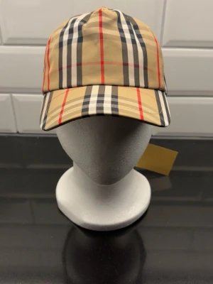 Beige keps med rutigt mönster Burberry - Säljer en klassisk beige keps från Burberry med det ikoniska rutiga mönstret i svart, vitt och rött. Kepsen har böjd skärm och justerbar rem bak. Tillverkad i ett lätt och bekvämt material som passar perfekt till streetwear och trendiga outfits.
