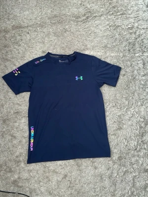 Mörkblå Under Armour t-shirt (reflex) - Snygg mörkblå t-shirt från Under Armour med regnbågsfärgade loggor och textdetaljer på bröstet, axeln och sidan. T-shirten har rund hals och är kortärmad, perfekt för en sportig och avslappnad stil. Det står att det är storlek M men passformen är som storlek S