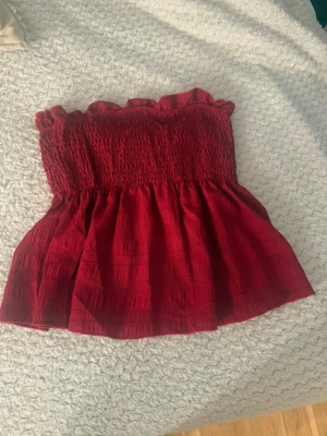 Röd bandeau topp med volangkant - Supersöt röd bandeau topp med smockad överdel och volangkant upptill. Toppen är axelbandslös och har en luftig, lätt utsvängd nederdel. Perfekt för varma dagar och enkel att styla med jeans eller kjol.