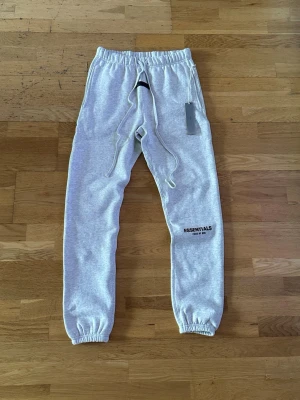 Grå mjukisbyxor från Essentials - Snygga ljusgrå mjukisbyxor från Essentials med tryck på benet. Byxorna har resår och dragsko i midjan samt muddar vid bensluten. Perfekta för en chill och avslappnad stil. Mjukt material som känns skönt mot huden. Det står storlek XS men passformen är som storlek S.