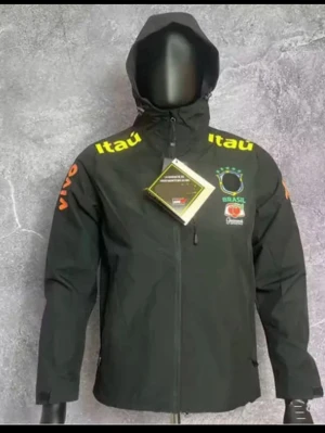 Svart Brasil Nike vindjacka med huva - Svart vindjacka från Nike med Brasil-logga och gula detaljer. Jackan har huva, dragkedja framtill, sidofickor och tryckta sponsorloggor som Itaú och Guaraná. Perfekt för dig som gillar sportig stil och vill visa stöd för Brasilien. Fleecefodrad