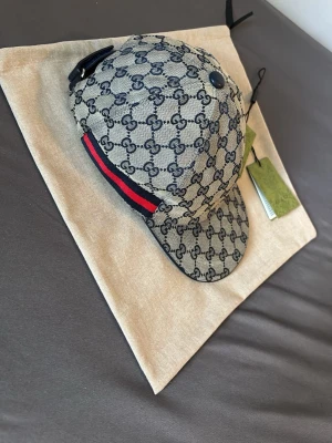 Gucci keps med GG-mönster och röd detalj - Snygg keps från Gucci i beige canvas med klassiskt GG-monogram över hela kepsen. Har en röd och marinblå rand på sidan och justerbar rem baktill. Perfekt accessoar för att lyfta din streetstyle. Kepsen har en böjd skärm och tydliga logodetaljer.
