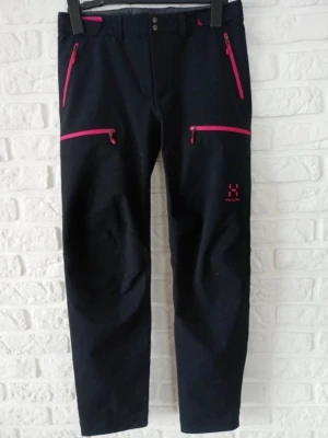Svarta byxor från Haglöfs med rosa detaljer - Svarta byxor från Haglöfs med snygga rosa dragkedjor och logga på benet. Byxorna har flera praktiska fickor med dragkedja och är perfekta för en aktiv livsstil. Materialet känns slitstarkt och bekvämt.
