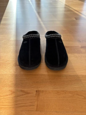 Svarta UGG tofflor i mocka - Säljer ett par svarta UGG tofflor i mjuk mocka med klassisk vit zigzag-detalj runt öppningen. Insidan är fodrad med fluffigt material för extra komfort. Perfekta att glida runt i hemma när du vill ha det extra mysigt.