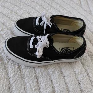 Svarta Vans sneakers,37 - Klassiska svarta Vans sneakers med vit sula och vita skosnören. Skorna har låg profil, canvasmaterial och tydliga vita sömmar. Perfekta för en avslappnad streetstil och passar till det mesta i garderoben. Storlek 37. Fint skick. 