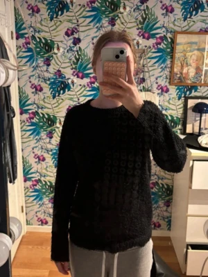 Svart fluffig stickad tröja - Mysig svart stickad tröja med fluffig textur och rund hals. Tröjan har långa ärmar och en enkel, rak passform som passar perfekt till både jeans och mjukisbyxor. Perfekt för kalla dagar när du vill vara både bekväm och stilren.