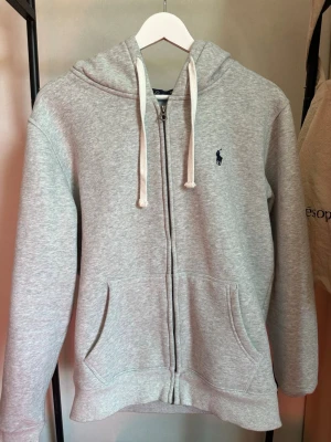 Grå zip-hoodie från Polo Ralph Lauren - Klassisk ljusgrå hoodie med dragkedja från Polo Ralph Lauren. Har huva med vita snören, två fickor fram och broderad marinblå logga på bröstet | Storlek: M | Använd fåtal gånger | Hör av er vid funderingar!