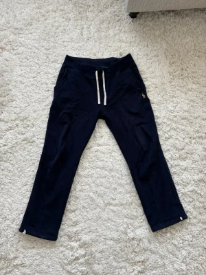 Mörkblå mjukisbyxor från Polo Ralph Lauren - Mörkblå mjukisbyxor från Polo Ralph Lauren med vit dragsko i midjan och klassisk gul logga på höften. Byxorna har raka ben och är tillverkade i mjukt bomullsmaterial som är skönt mot huden. Perfekta för chill dagar hemma eller när du vill vara bekväm.