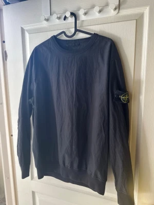 Svart  tröja från Stone Island - Snygg svart stickad tröja från Stone Island med klassisk rund hals och ribbade muddar. Tröjan har den ikoniska logotypen på vänster ärm och är perfekt för dig som gillar stilrena och trendiga plagg.