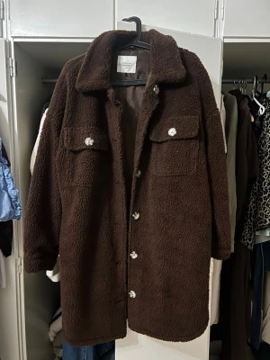 Brun teddyjacka från Jacqueline de Yong - Mysig brun teddyjacka från Jacqueline de Yong med stora bröstfickor, knäppning framtill och klassisk krage. Jackan är tillverkad i ett fluffigt teddy-material som håller dig varm och har en avslappnad passform. Perfekt för kyliga dagar.