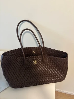 Mörkbrun flätad shoppingväska från Nelly - Snygg mörkbrun shoppingväska i flätat konstläder med dubbla handtag och guldfärgad detalj framtill. Väskan har en rymlig form och stängs med tryckknapp. Perfekt för dig som vill ha en trendig och praktisk väska med stilren look.