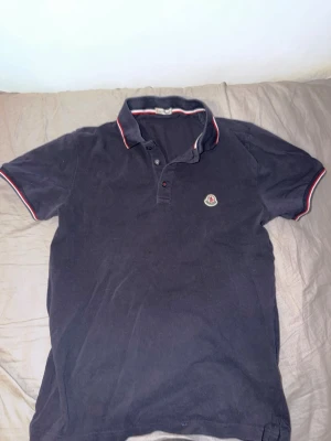 Moncler Piké  - Mörkblå pikétröja från Moncler med klassisk krage och knäppning framtill. Tröjan har röda och vita detaljer på krage och ärmslut samt Moncler-logga på bröstet. Perfekt för en stilren och sportig look.