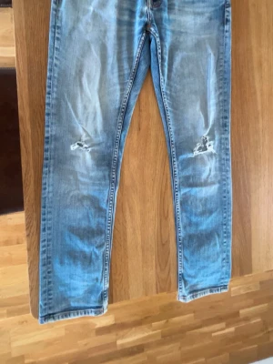 Nudie Jeans - Säljer ett par ljusblå jeans från Nudie Jeans med slitningar och hål på båda knäna. Klassisk femficksmodell med raka ben och snygg tvätt. Jeansen har kontrastsömmar och är tillverkade i bomull. Perfekta för en avslappnad och trendig look. Dom är strlk 32 30