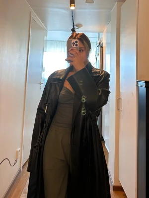 Svart lång trenchcoat i skinnimitation - Snygg svart trenchcoat i skinnimitation med bälte i midjan och klassisk krage. Jackan har långa ärmar, knäppning framtill och coola detaljer som axelklaffar och fickor. Perfekt för dig som vill ha en edgy och trendig look.