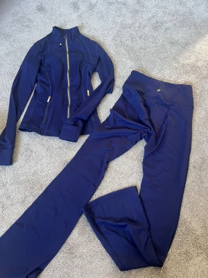 Mörkblått träningsset från Lululemon - Storlek 2, Snyggt mörkblått träningsset från Lululemon med figurnära jacka med dragkedja och långa ärmar samt matchande utsvängda tights. Materialet är stretchigt och följsamt, perfekt för träning eller yoga. Jackan har hög krage och diskreta logodetaljer.