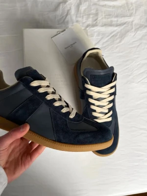 Maison Margiela Gats - Snygga mörkblå sneakers från Maison Margiela med ovandel i mocka och läder, beigea skosnören och klassisk gummisula. Perfekt för dig som gillar stilrena och tidlösa sneakers med en lyxig känsla. Diskret logga på plösen och låg modell.