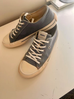 Ljusblå sneakers från Axel Arigato - Snygga låga sneakers från Axel Arigato i ljusblå canvas med vita detaljer och gummisula. Klassisk design med vita snören och tåhätta. Perfekta för en stilren och avslappnad look. Små fläckar på höger skon men det går fixa med textil penna eller tjockare snören 
