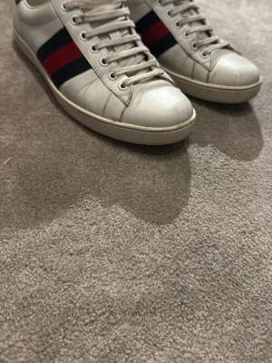 Gucci vita sneakers med röd/blå rand - Snygga vita sneakers från Gucci med klassisk röd och blå rand på sidan och mörkblå häl i ormskinnsmönster. Skorna har vita snören och låg profil, perfekt för en trendig streetstyle-look.