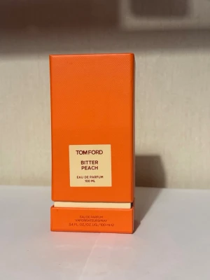 Tom Ford Bitter Peach EdP 100 ml - Tom Ford Bitter Peach Eau de Parfum i en snygg orange glasflaska på 100 ml. Parfymen kommer i en matchande orange kartong med stilren design. Flaskan har en fyrkantig form och tydlig etikett framtill. Perfekt för dig som gillar lyxiga och fruktiga dofter.