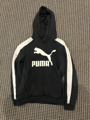 Svart Puma hoodie med vita ränder - Svart hoodie från Puma med stor vit logga och text på bröstet. Tröjan har huva och vita ränder längs ärmarna. Tillverkad i mjukt material som känns skönt och sportigt. Perfekt för dig som gillar streetwear och sportig stil.