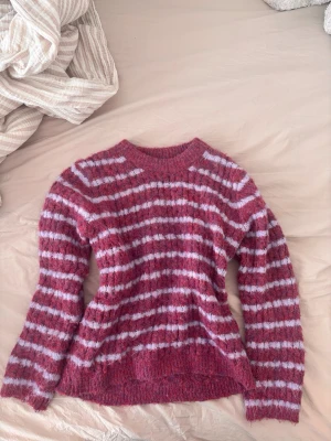 Rosa-lila-randig stickad tröja fr Magasin du Nord - Mysig stickad tröja från Magasin du Nord i rosa med syrenlila horisontella ränder. Tröjan har en fluffig, lite luddig textur och rund halsringning. Perfekt för kalla dagar och enkel att styla med jeans eller kjol. Köpt för runt 800kr i Danmark 
