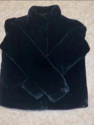 Svart fluffig pälsjacka - Mysig svart pälsjacka med hög krage och rak passform. Jackan har långa ärmar och är tillverkad i ett mjukt, fluffigt material som ger en lyxig känsla. Perfekt för kalla vinterdagar när du vill hålla dig varm och snygg.