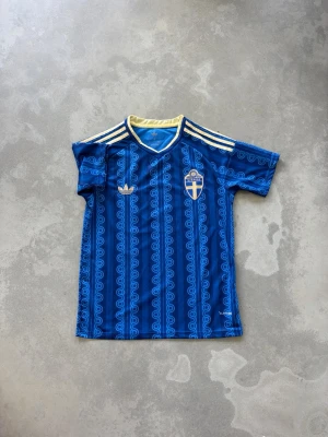 Sveriges bortaställ 2026 - Blå fotbollströja med Sveriges landslagsemblem på bröstet. Tröjan har gula detaljer, vita Adidas-ränder på axlarna och ett unikt mönster över hela tyget. Perfekt för dig som vill visa ditt stöd för Sverige på matchen eller träningen.