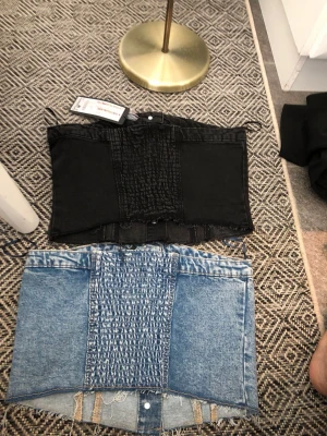 Två denim tubtoppar med knappar - Två coola tubtoppar i denim, en i svart och en i ljusblå tvätt. Båda har knappar framtill, råa kanter och smockad detalj bak. Snygga fickor framtill och en edgy, croppad passform. Perfekta för att lyfta din outfit med en unik touch.