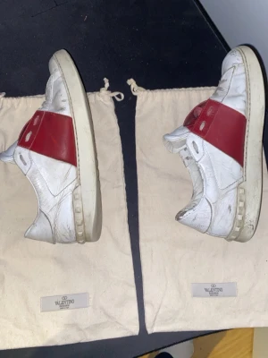 Valentino vita sneakers med röd detalj - Säljer ett par vita sneakers från Valentino storlek 42 med en bred röd läderdetalj över sidan. Skorna har klassisk låg profil, vita snören och sula i offwhite. Tillverkade i läder och har en stilren, sportig look med subtila logodetaljer. Köpta på StoreEnda 100% äkta! Box, verifikations kort, dustbags och digitalt kvitto medföljer. !OBS skorna sitter mer som 43 än 42.                 (Kan gå ner i pris vid snabb affär 
