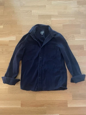 Mörkblå overshirt från H&M - Stilren mörkblå overshirt från H&M i regular fit. Skjortan har två bröstfickor med lock, knäppning framtill och klassisk krage. Perfekt att slänga över en t-shirt för en avslappnad look.