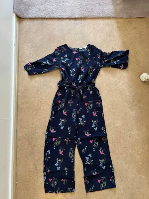 Mörkblå blommig jumpsuit med knytskärp - Snygg mörkblå jumpsuit med blommigt mönster i rosa, blått och grönt. Modellen har långa ärmar och ett knytskärp i midjan. Vida ben och mjukt, lätt tyg som ger en avslappnad look.