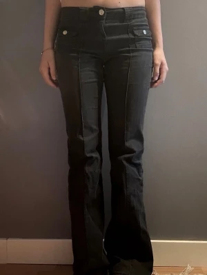 Svarta bootcut jeans med fickdetaljer - Snygga svarta bootcut jeans med markerade sömmar och fickor framtill med lock och silverknappar. Hög midja och utsvängda ben ger en trendig siluett. Perfekta för dig som gillar en retrovibe och vill sticka ut lite extra.