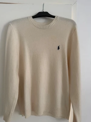 Vit stickad tröja från Polo Ralph Lauren - Klassisk vit stickad tröja från Polo Ralph Lauren med rund halsringning och ribbade muddar. Tröjan har den ikoniska mörkblå loggan broderad på bröstet och är tillverkad i mjukt material som känns lyxigt mot huden. Tröjan är nästan helt ny. Använd 2 gånger, den är lite för stor för mig. Nypris 2499. 