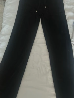  svarta mjukisbyxor med fickor - Svarta mjuk byxor med raka ben och dragsko i midjan. Baks XSidan channel två stora fickor med silverfärgade knappar. Perfekta för chill eller streetwear. Materialet ser ut att vara mjukt och stretchigt, troligen bomull eller bomullsblandning.