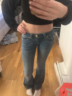 Blå bootcut jeans True Religion - Säljer ett par blå bootcut jeans från True Religion med låg midja, kontrastsömmar och stora dekorativa knappar på bakfickorna. Jeansen har klassisk logga på etiketten bak och är gjorda i denim. Perfekt för dig som gillar en trendig och avslappnad look.